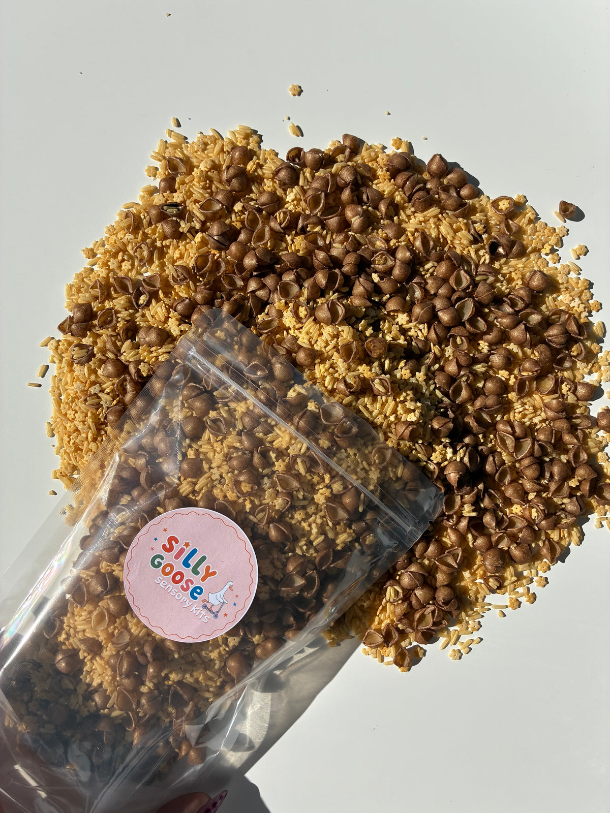 A clear stand-up pouch with a round pink 'Silly Goose' sticker, filled with dried pasta and rice. Sensory filler titled 'Cookie Crumble'